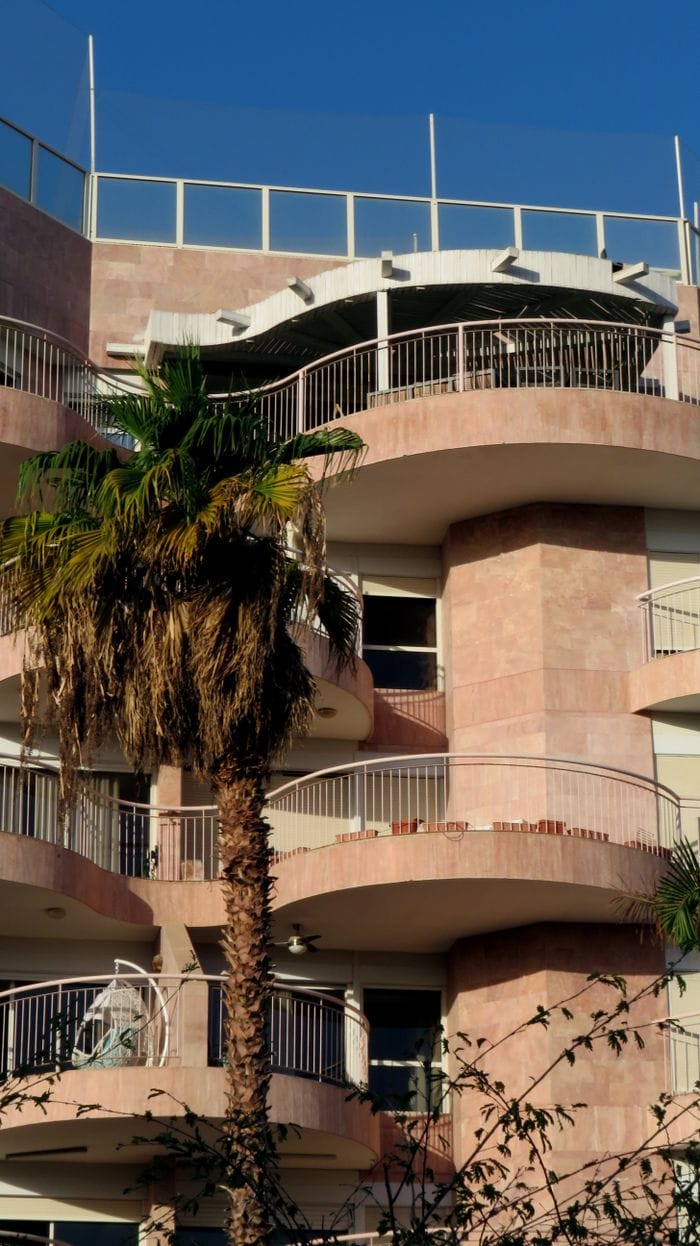 our-story-02 Contemporary urban apartment building with palm trees against blue sky.